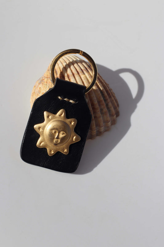 Black leather keychain with gold sun design on a shell against a light gray background