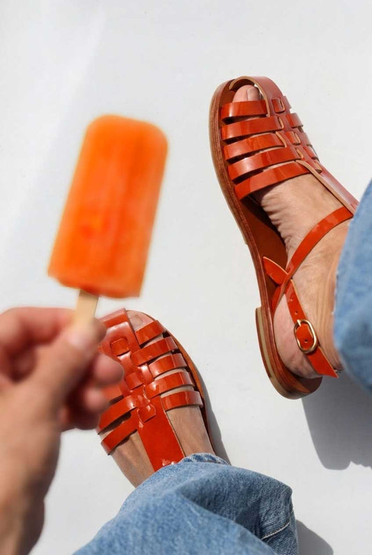 Person wearing orange sandals holding an orange popsicle against a white background