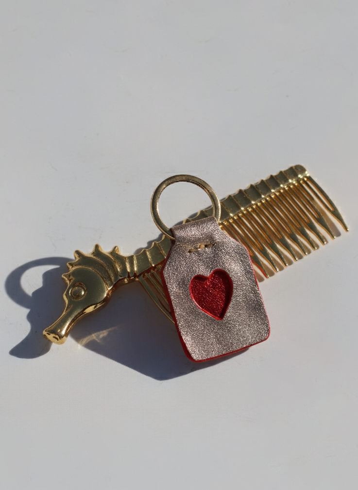 Gold comb charm with a red heart tag on a light gray background