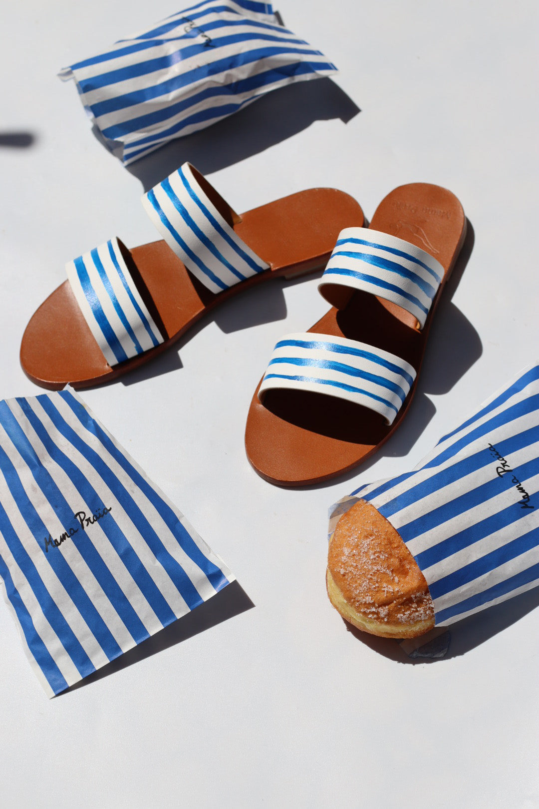 Brown sandals with blue striped straps on a white surface with blue striped towels and a pastry.